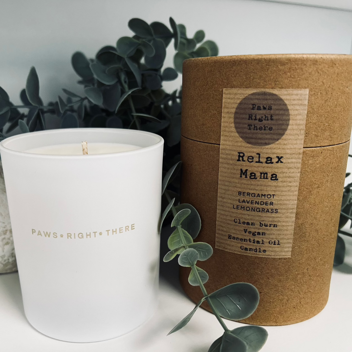 Candle in a matt white jar, engraved with Paws Right There. It sits next to a kraft brown cardboard tube labelled Relax Mama. this tube is the packaging for the candle